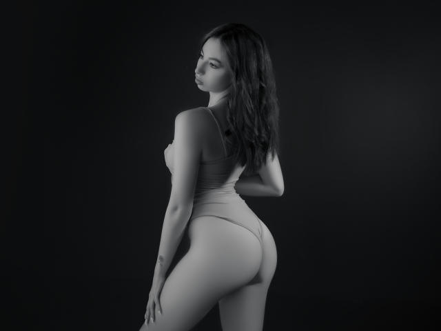Camille Rios, con lunghi capelli scuri, indossa un costume intero grigio aderente che mette in mostra il sedere e i piccoli seni, mentre si volta a guardare, in piedi contro uno sfondo scuro, in una fotografia in bianco e nero..