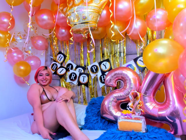 Charlize Addams con cabello rosa, grandes senos en un bikini marrón brillante, sentada en una cama con pelaje azul, rodeada de globos amarillos y rosas, y un globo brillante con el número "4"..