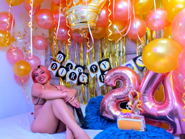 Charlize Addams con cabello rosa, desnuda, sentada en una cama con las piernas cruzadas, rodeada de globos naranjas y amarillos, un globo rosa con el número "4", y un cojín azul y esponjoso..