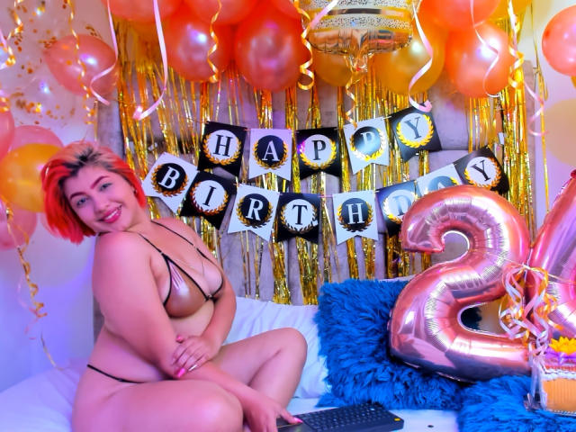 CharlizeAddams con i capelli rossi, che indossa uno slip nero e una canottiera nera, mostrando il suo seno prosperoso, seduta su un letto con cuscini blu, circondata da palloncini e un palloncino con il numero "2"..