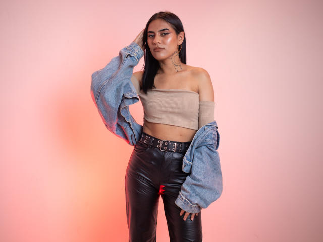 MeganXBrown tem cabelo preto, uma blusa marrom com um ombro à mostra, calças de couro pretas e uma jaqueta jeans azul. Ela está em pé com uma mão na cabeça, parecendo confiante..