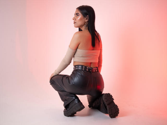 MeganXBrown, kneeling, wearing a tan off-shoulder top, black leather pants, and black boots, showing her ass, with long black hair and earrings, under pink lighting.