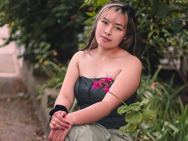 SallyDoll with medium tits, wearing a strapless black top with pink flower, green skirt, black wristband, sitting in garden, looking at camera, blonde hair, slightly chubby.