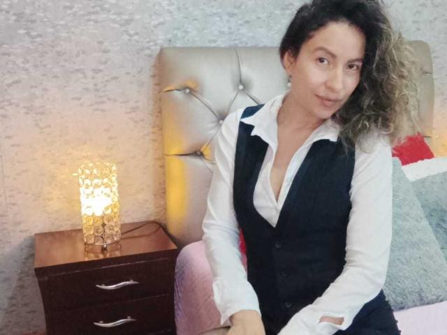 EroticSasha, mit lockigem, braunem Haar, sitzt auf einem Bett, trägt ein weißes Hemd und eine schwarze Weste und lächelt leicht. Eine Lampe und ein Nachttisch aus dunklem Holz befinden sich links..