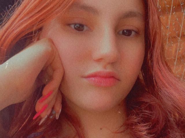 EstefaniaRamos: Ze heeft rozeachtige rode haren, roze lippenstift en roze nagels, en leunt haar wang op haar hand, met een licht verveelde blik en halfgesloten ogen..