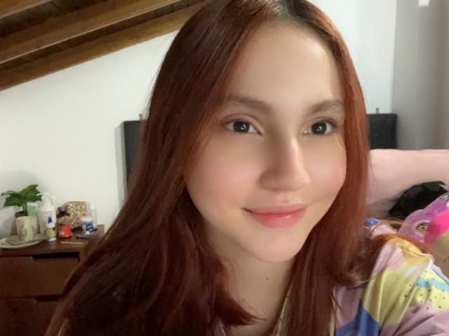 EstefaniaRamosHot with red hair, light skin, brown eyes, and pink lips, smiling softly. She's wearing a colorful shirt, and her boobs are partially visible.