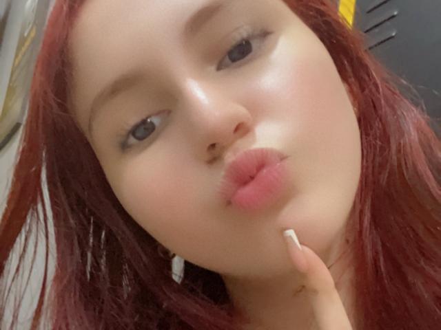 EstefaniaRamos, bellissima, con labbra rosa gonfie, capelli rossi e pelle chiara, che mostra la lingua e tocca il mento con un dito con unghie curate..
