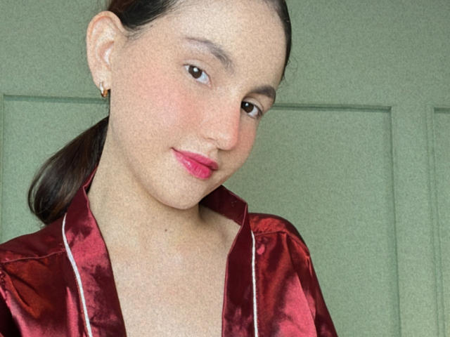 EstefaniaRamosHot con lápiz labial rosa, ojos marrones y cabello castaño recogido en una coleta, vistiendo una camisa roja de satén brillante, pequeños aretes dorados y una ligera sonrisa..