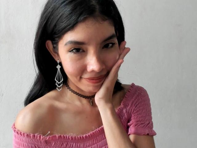MileyWatson with black hair, pink off-shoulder top, choker, and dangling earrings, smiling, hand on cheek, showing cute tits, soft skin, subtle makeup, gray background.