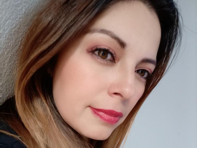 Seher's face with brown eyes, pink eyeliner, red lipstick, and light brown, slightly wavy hair, smiling softly. Her skin is pale, and she's wearing a black top.