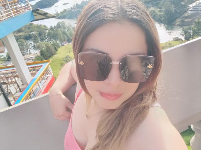 Seher with brown hair, wearing pink top, brown sunglasses with gold star patterns, smiling, holding her top strap, standing on a balcony, showing her tits.