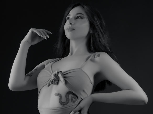 Black-and-white photo of CamilleRios with long dark hair, small tits, tattoo of a snake on her lower right rib, wearing a tied-up bra, looking up with a confident expression.