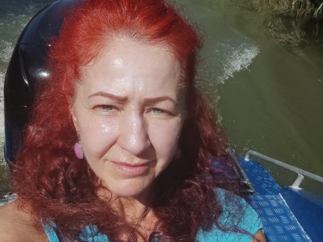 MilayaDFlow with red curly hair, sweaty face, and pink earrings, sitting in a blue boat, smiling slightly, wearing a blue top, water splashing around.