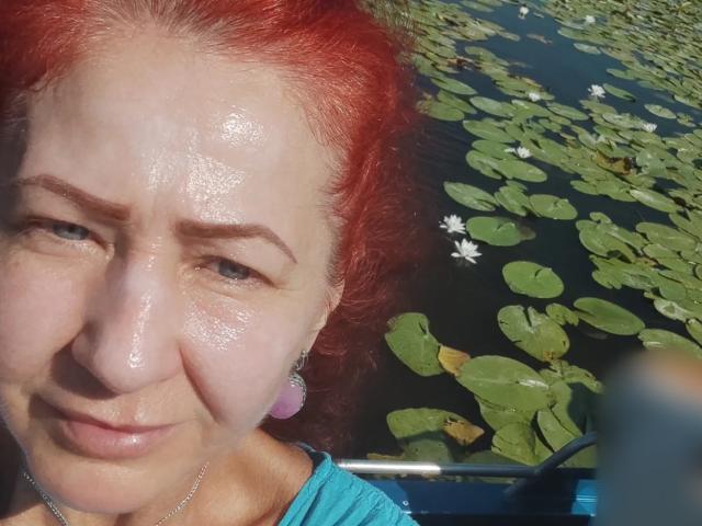 MilayaDFlow met glanzend rood haar, een nat gezicht, kleine roze oorbellen, een blauw shirt, die lichtjes glimlacht, drijvend op een boot over water bedekt met lelies, met kleine witte bloemen..