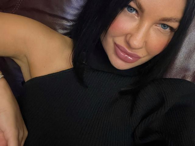 KristenStark with blue eyes, black hair, pink glossy lips, wearing a black ribbed sleeveless top, sitting on a dark brown leather couch, smiling at the camera.
