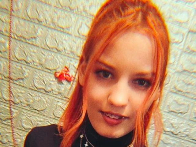Love69Hottie has red hair, a slight smile, and is wearing a black turtleneck. She has a small nose and visible metal teeth. The image is slightly blurry.