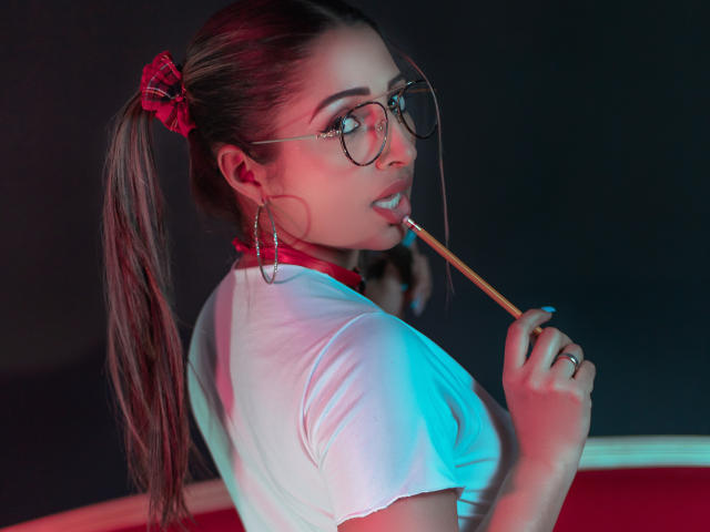 StefanyCaruso, avec des lunettes, un élastique pour cheveux rouge à carreaux, un t-shirt blanc, un col rouge, tenant un crayon près de sa bouche, des boucles d'oreilles cerceaux, une queue de cheval sur le côté, un éclairage bleu et rouge, regardant en arrière..