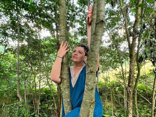 Carolina69 gripping a tree with one hand, wearing a blue dress, looking up with a slight smile, surrounded by green foliage. She's holding onto a branch with her other hand.