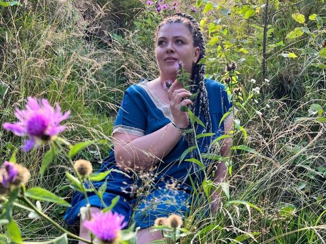 Carolina69 with braided hair, blue dress, and white lace, holding a flower, sitting in a grassy field with purple and yellow flowers in the foreground.