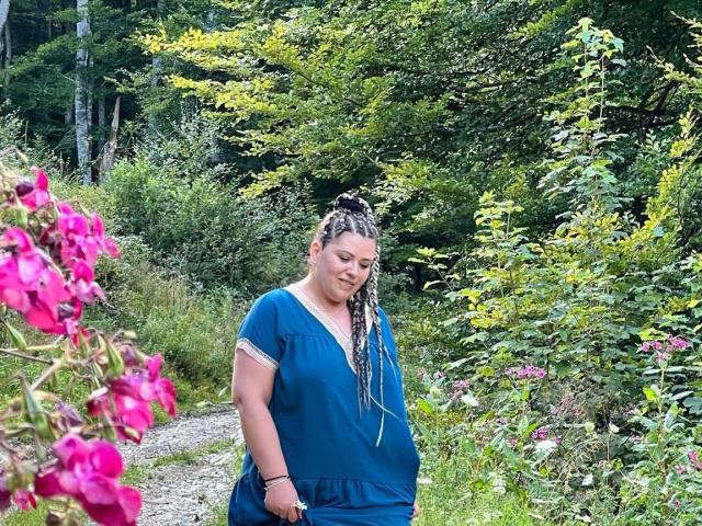 Carolina69, chubby with long blonde braids, wears a blue dress, standing on a gravel path, surrounded by greenery and pink flowers, looking down with a slight smile.