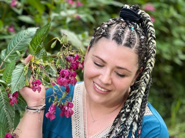 Carolina69 with braided hair, blue shirt, and white lace, smiling while holding pink flowers. She's holding a bunch of flowers in her right hand, looking down.