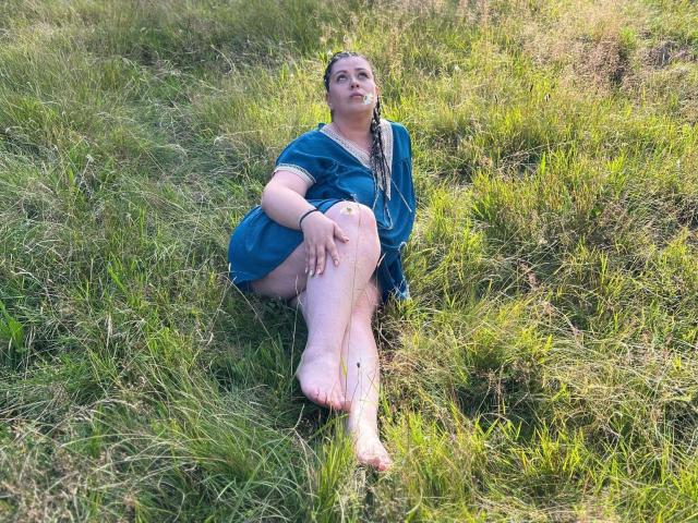 Carolina69 with braided hair, blue dress, and bare feet, sitting in grass, looking up, legs crossed, left knee up, right knee down, green grass all around.
