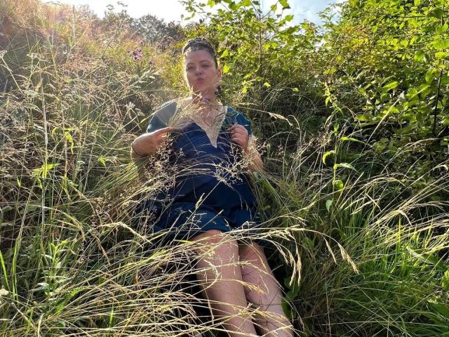 Carolina69 is in a grassy field, holding tall grass, wearing a blue dress, with her ass partially covered, looking slightly to the side, in sunlight.