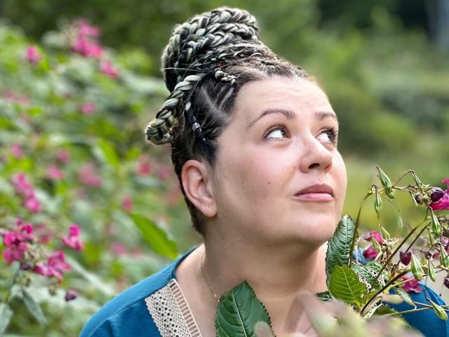 Carolina69 with braided updo, looking up with a curious expression, holding pink flowers, wearing a blue dress with lace, green leaves in the foreground, blurred background.