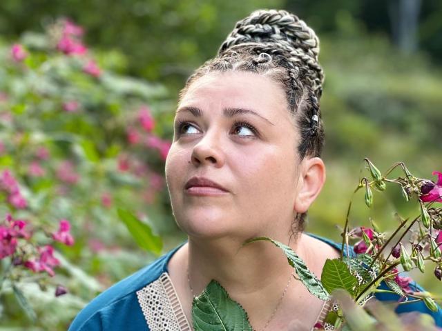 Carolina69 with braided hair, blue shirt, and green leaves, looking up with a slight smile, her ass and tits partly visible, surrounded by pink flowers and greenery.