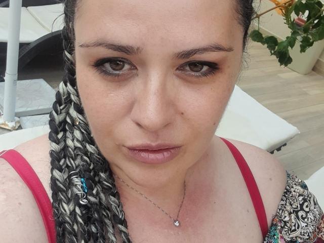 Carolina69 with puffy lips, brown eyes, and braided black and white hair. She's wearing a red top and a patterned dress, looking straight at the camera.