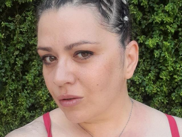 Carolina69 has light skin, dark braided hair, small brown eyes, subtle makeup, and a red tank top. Her face shows a slight pout with small tits visible.