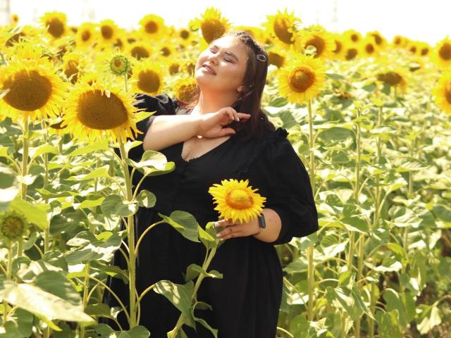 KaidoKun met gele zonnebloemen, waarbij ze er één in haar rechterhand houdt, gekleed in een zwarte jurk, met gesloten ogen, bruin haar, zonlicht, groene bladeren, lange stelen, zomerweide, kalme uitdrukking..