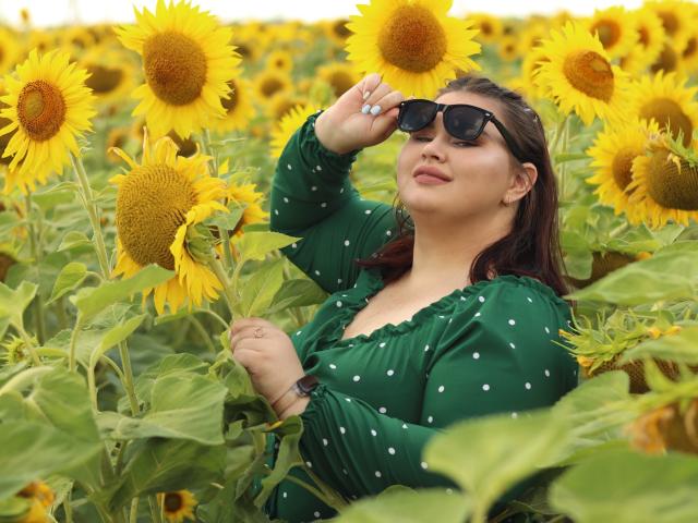 KaidoKun with sun-kissed skin, wearing dark sunglasses, green polka dot dress, holding sunflowers, eyes closed, smiling, brown hair, standing in sunflower field, sunny day.