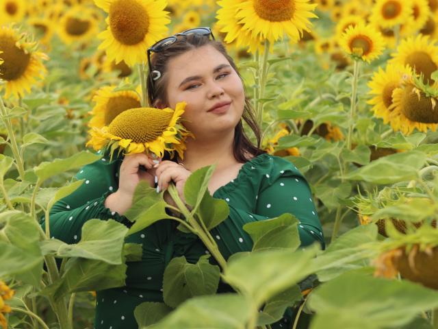 KaidoKun met gele zonnebloemen, een zonnebloem vasthoudend, groene jurk met stippen, zonnebril op het hoofd, lachend, middelgrote borsten, bruin haar, in een zonnebloemenveld, zomer, blij..