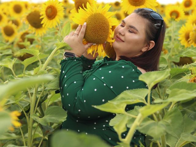KaidoKun met een door de zon gekleurd gezicht, houdt een gele zonnebloem voor zijn neus, draagt een groene jurk met stippen, heeft bruin haar, draagt een zonnebril op zijn hoofd, glimlacht en staat in een veld met zonnebloemen, en ziet er blij uit..