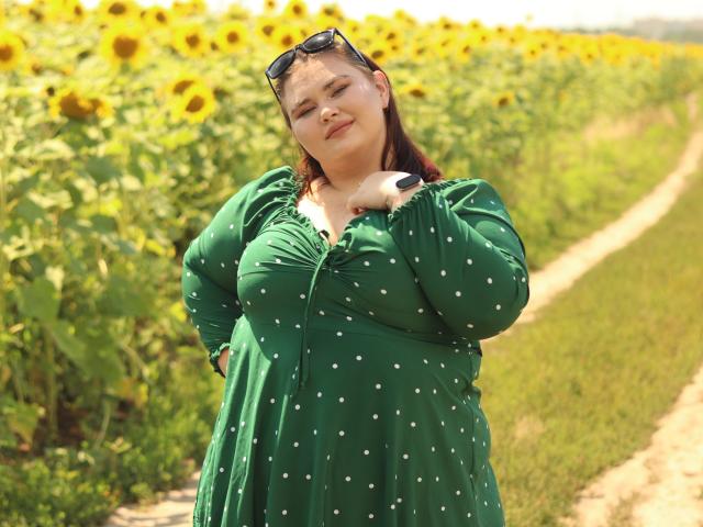 KaidoKun, a chubby woman with large tits, green polka dot dress, hand on chest, sunflowers, dirt path, sunglasses on head, smiling, sunlight, summer, relaxed pose.