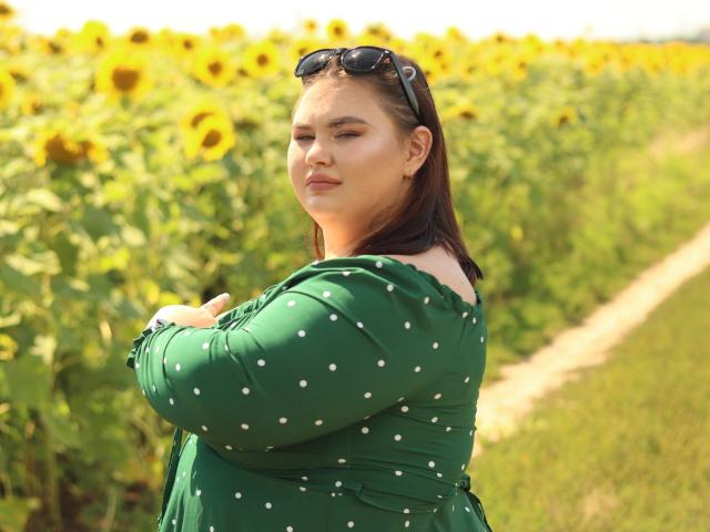 KaidoKun porte une robe verte à pois, a les bras croisés et porte des lunettes de soleil sur la tête, se tenant dans un champ de tournesols, avec un léger sourire et des cheveux bruns..