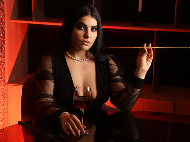 Melissa, avec des cheveux noirs, une robe noire transparente, un collier de perles, tenant une baguette, un verre de vin rouge, assise, éclairage rouge, expression sérieuse, poitrine généreuse, table en verre, fond sombre..
