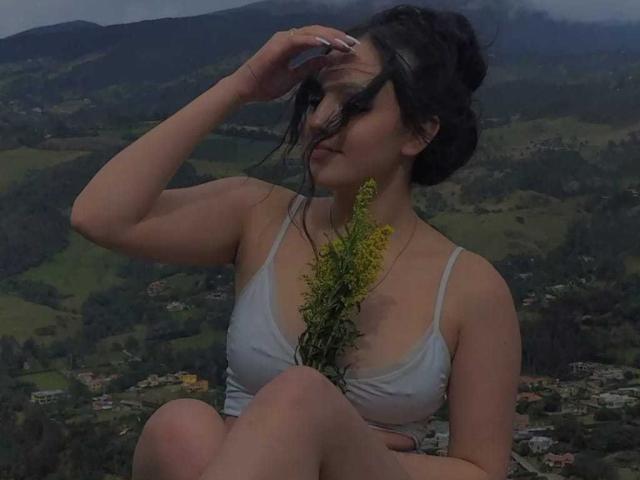 HaleiBeckham com cabelo escuro em um coque bagunçado, segurando flores amarelas, vestindo uma camiseta branca, sentada com os joelhos levantados, mão no cabelo, ao ar livre, céu nublado..