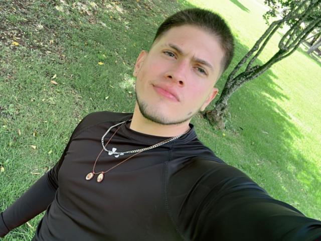 SebastiianMaxime, avec ses cheveux bruns courts, sa peau claire et sa légère barbe, portant une chemise noire et deux pendentifs, prend un selfie en extérieur, souriant légèrement..