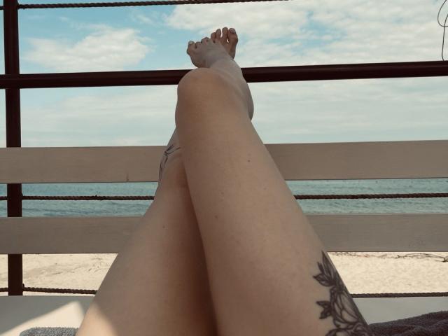Tsukerberg's legs crossed at the ankles, showing tattooed thighs, with feet up, resting on a wooden bench, against a beach and ocean view, under a blue sky.