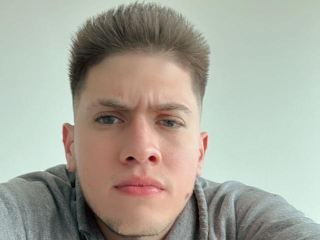 SebastiianMaxime, avec une expression sérieuse, légèrement agacée, des cheveux bruns courts et hérissés, des yeux bleus et une chemise grise. Son nez est légèrement rouge et ses sourcils sont froncés..