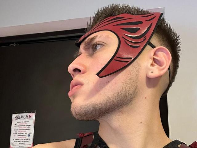 SebastiianMaxime indossa una maschera rossa e nera che gli copre gli occhi, ha i capelli marroni a punte e una barba. Sta guardando di lato, mostrando il naso e l'orecchio..