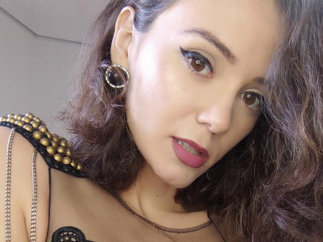 PrettyWanda con i capelli castano ricci, che indossa un rossetto rosso acceso, una maglia nera in pizzo con borchie dorate e orecchini a cerchio, guarda la fotocamera con un leggero sorriso..