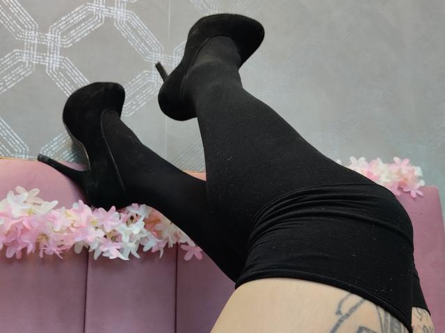 De benen van AmySpider, bedekt met zwarte, glinsterende leggings, zwarte hoge hakken, en een roze bank bekleed met bloemen. Een zichtbare tatoeage op de rechter dij..