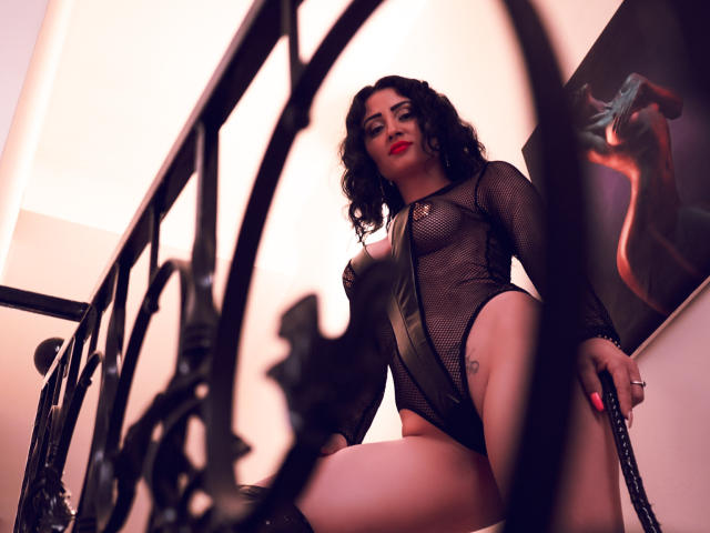 KarinaGray, avec ses cheveux noirs bouclés, portant une combinaison noire en mesh et en cuir, qui révèle ses seins et ses parties intimes, se tient sur un escalier, tenant un fouet, et regarde en bas de manière séduisante..