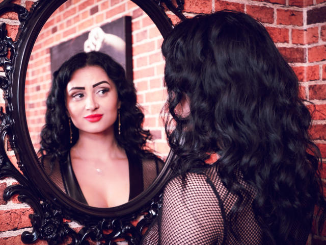 Karina Gray, con cabello negro ondulado, lápiz labial rojo y top de rejilla, mirando su reflejo en un espejo ornamentado, mostrando sus senos en la imagen. Fondo de pared de ladrillo..