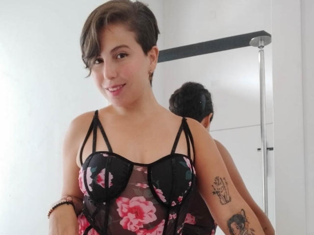 Alice Howard, con i capelli castani corti, indossa un abito intimo nero con fiori e si sta guardando allo specchio, mostrando il seno. Ha dei tatuaggi sul braccio e sta sorridendo..