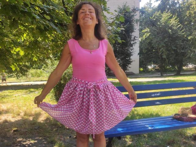 GrostFlow está en un parque soleado, vestida con una camiseta rosa y una falda rosa con estampado floral, sosteniendo el dobladillo de la falda, sonriendo con el pelo castaño rizado y la piel bronceada..