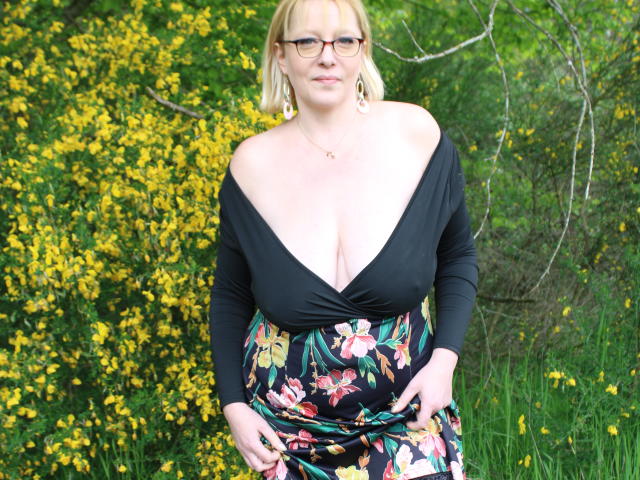 EleanaRose ha un seno prosperoso, indossa una maglia nera a spalle scoperte e una gonna floreale, ed è in piedi con dei fiori gialli sullo sfondo, indossando occhiali e orecchini dorati..